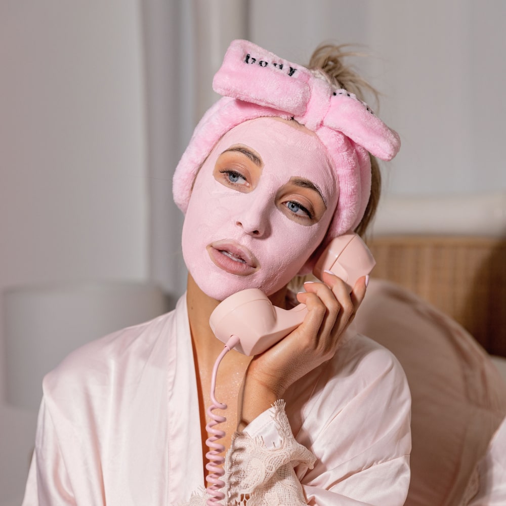Woman wearing a pink skincare face mask and BodyBlendz pink bow headband while holding a phone, relaxing during self-care routine at home